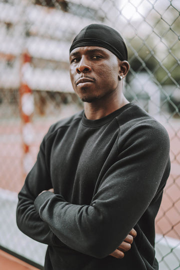 A man in the street in front of a fence wearing a durag.